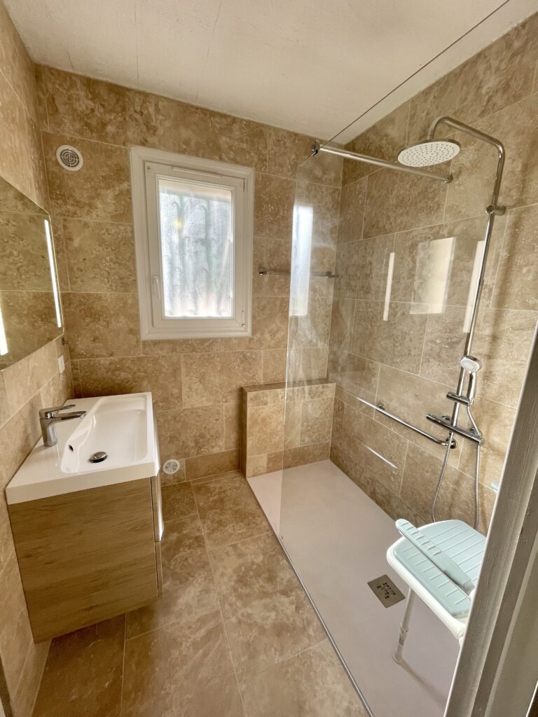 Salle de bain : carrelage beige, lavabo blanc, fenêtre givrée, douche en verre avec pommeau de douche à effet pluie et chaise blanche