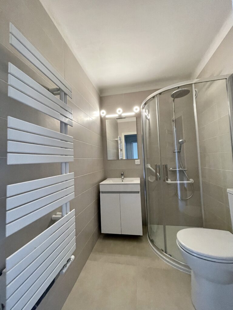 Salle de bain : carrelage beige, douche en verre bombé, radiateur mural, vasque et miroir blancs, toilettes.