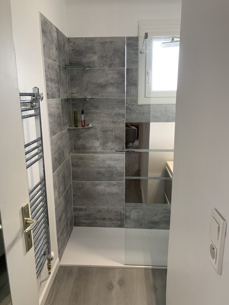Salle de bain moderne : douche italienne, murs gris, porte-serviettes argentées, petite fenêtre.