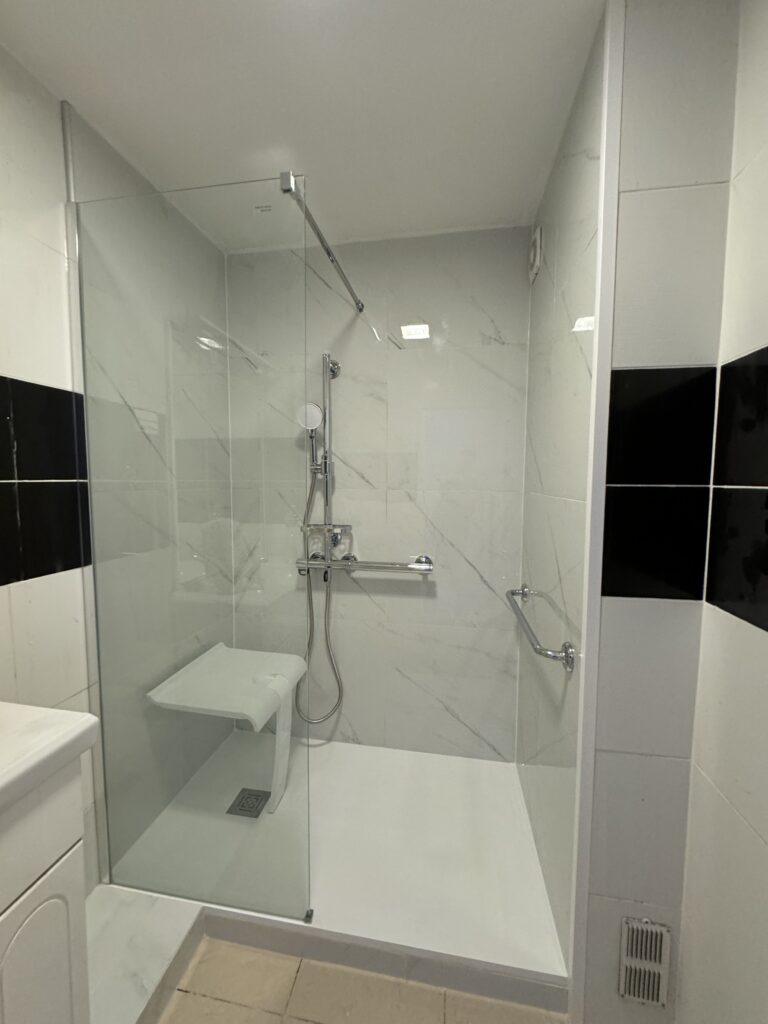 Douche PMR moderne avec siège intégré, barres de soutien, carrelage blanc et accents noirs.