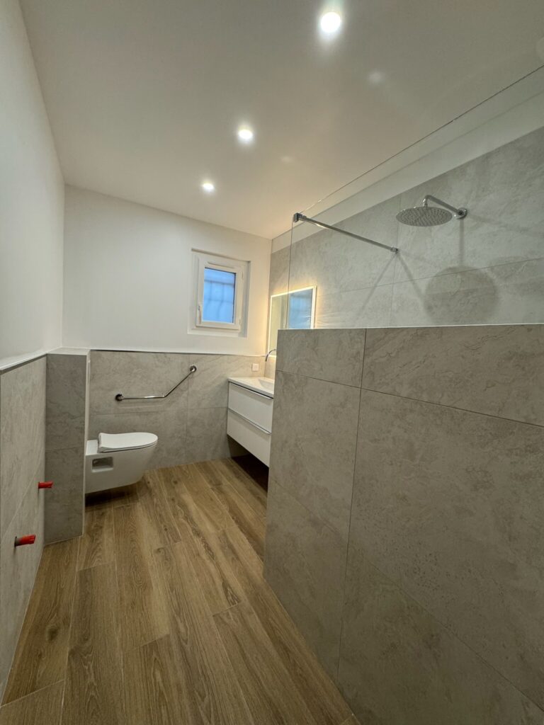 Salle de bains moderne : WC suspendu, grande douche, sol bois.