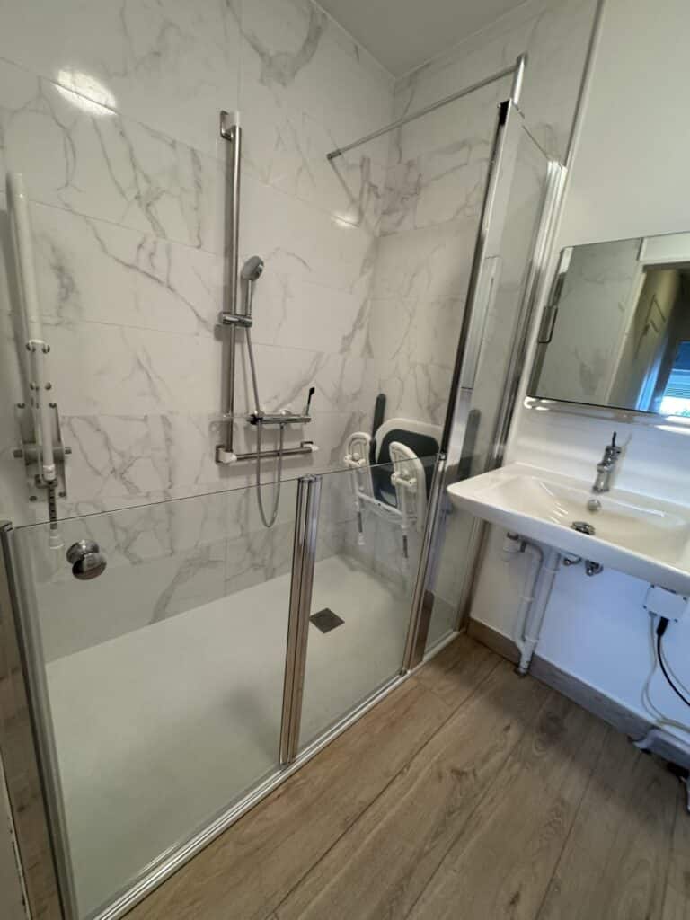 Salle de bain PMR rénovée : douche italienne, barres d'appui, siège et lavabo suspendu.