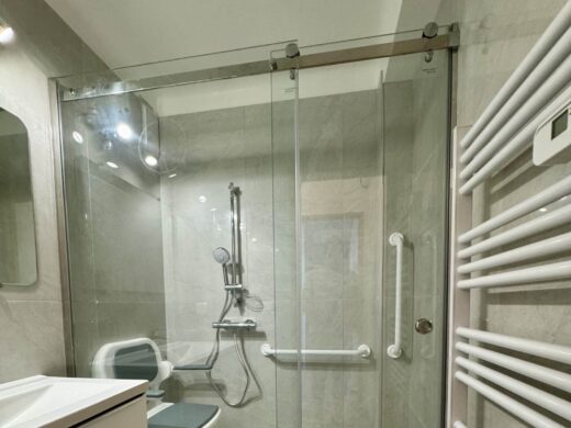 Salle de bain moderne avec douche vitrée, siège et barres de soutien.